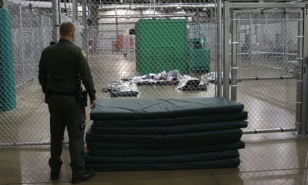 Man on one side of cyclone gate watches people sleeping on cement floor under bright lights.