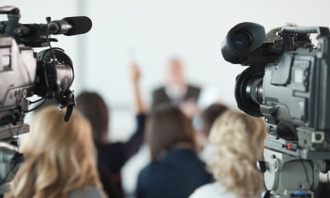 Cameras pointed at a blurry image of a person giving a presentation with mostly women reporters in the midground