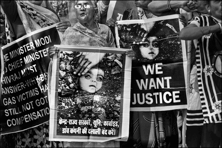 As mulheres seguram cartazes que mostram o rosto de uma criança semienterrada, com a mão de um adulto limpando a sujeira do cadáver.  Um cartaz diz 'Queremos justiça';  outro diz 'O primeiro-ministro Modi e o ministro-chefe Shivraj devem responder por que as vítimas do gás de Bhopal ainda não receberam compensação.'