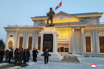 Kim Jong-un in front of a museum with a statue and a flag and several officials in black