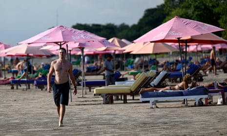 A man walks along a beach in Bali while other tourists lie on sun chairs under umbrellas