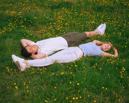 A man and a woman lie in grass studded with buttercups, wearing t-shirts