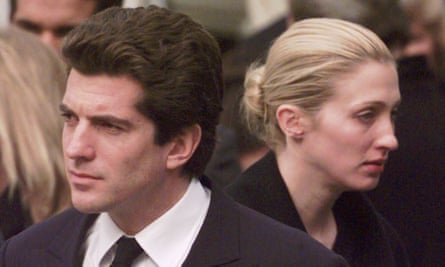 A young white man in a suit standing in front of a young white woman with blond hair pulled back into a bun. Neither is smiling.