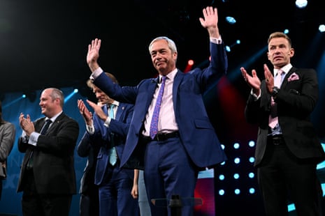 Nigel Farage in a conference venue, standing with both hands raised while Reform members applaud behind him