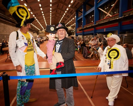 Three men in white suits and oversized rosettes. One is holding a ventriloquist dummy