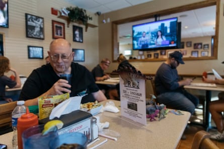 a man grins in a restaurant at a table with a few plastic dinosaurs and a sign