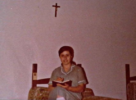 A young Claudia Carrero sitting holding a book with a small crucifix on the white wall behind her