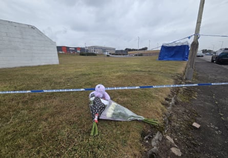 Floral tributes and a pink teddy bear lie behind police tape at the scene