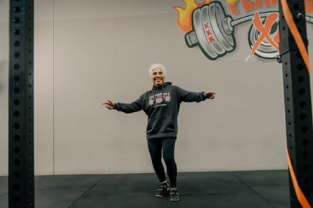 A woman with short white hair wearing black leggings and a dark-grey hoodie, smiling and standing with her arms outstretched in a gym