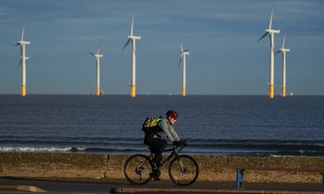Man rides a bike on a road by the sea with offshore windfarm in the background.