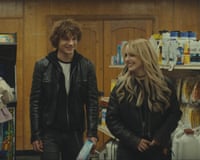 Two teenagers wearing leather jackets smile at each other inside a convenience store