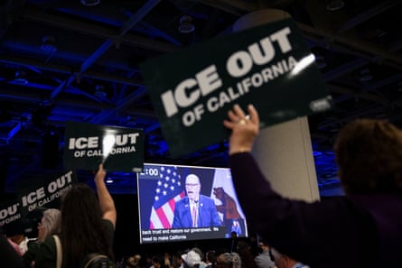 people hold up black and white signs saying ‘ICE out of california’ as a man in suit speaks on stage