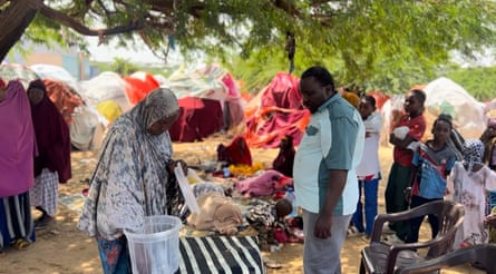 A woman stands at a table under a tree while a man looks on. Next to them are a group of children, and behind them several women sleep on a mat while in the background tents covered with bright fabric can be seen.