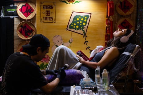 Tattoo artist works on the thigh of someone wearing headphones, holding an iPhone and reclining back in a chair