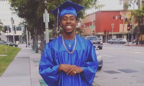 A man stands on a city street wearing a blue graduation cap and gown.