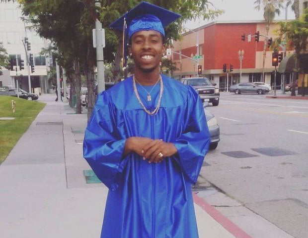A young man wearing dark blue graduation robes poses on the street.