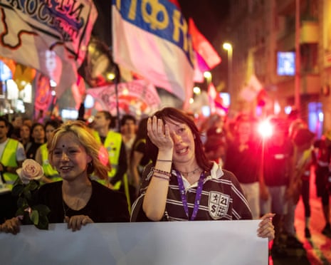 A student wipes her eye amid a crowded night march with flags and a red flare, Belgrade