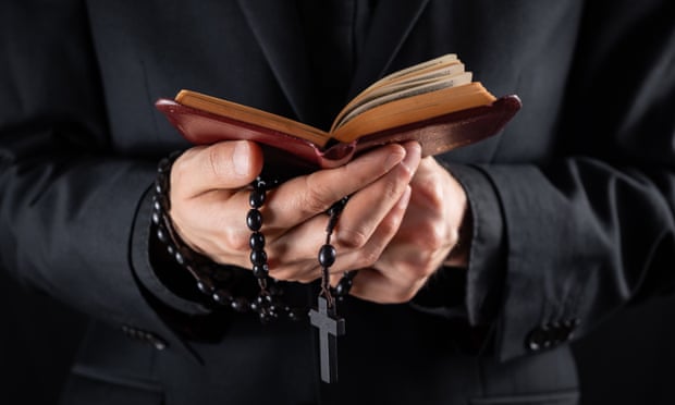Hands of a christian priest dressed in black holding a crucifix and reading New Testament book.