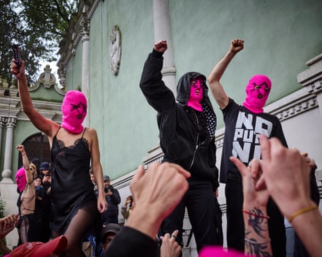 Women in pink balaclavas raise their fists in the air as they protest outside a green-coloured building