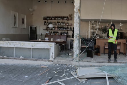 A man in hi-vis waistcoat looks at his mobile phone while standing alongside the blown-out frontage of the restaurant. Inside, wine bottles still stand on shelves