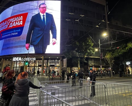 Police guard barricades in Belgrade as a screen shows leader Aleksandar Vucic on election night
