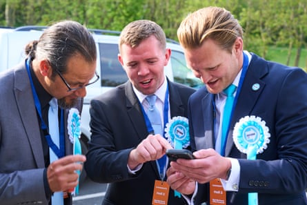 Three men in Reform rosettes and blue suits look at a phone