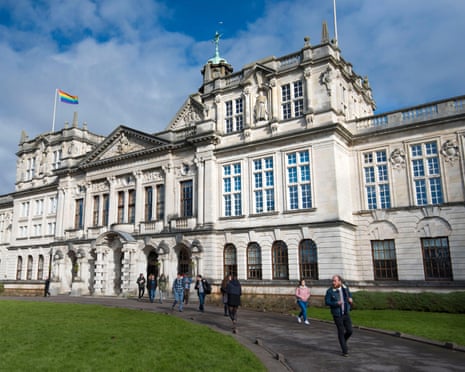 The main Cardiff university building, completed in 1909: it is pale stone with arched windows on the ground floor and pillars around a grand entrance. Students walk along a path in front.