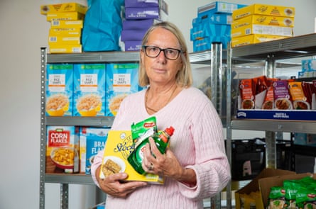 Tina McGrath poses in front of shelves of groceries at the food bank. She is holding a few items including washing-up liquid and wheat cereal biscuits