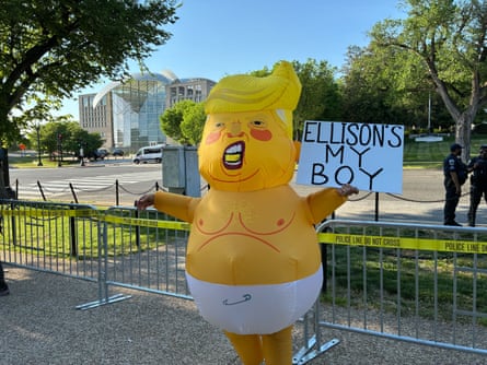 A yellow blow-up costume of Donald Trump in a diaper, with the person inside holding a sign that says ‘Ellison’s My Boy.’