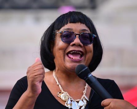 Diane Abbott wearing sunglasses speaks into a microphone, raising a fist as she addresses a crowd.