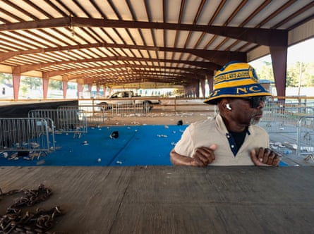 Man wearing yellow and blue bucket hat, standing under a ribbed roof that covers a truck and trash-strewn ground in the distance