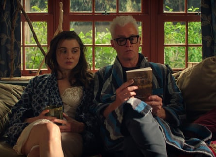 Rachel Weisz and John Slattery sitting side by side on a sofa wearing dressing gowns, while he reads the back cover of a book