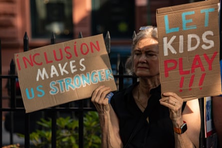 woman stands outside holding two signs saying ‘inclusion makes us stronger’ and ‘let kids play!!!’