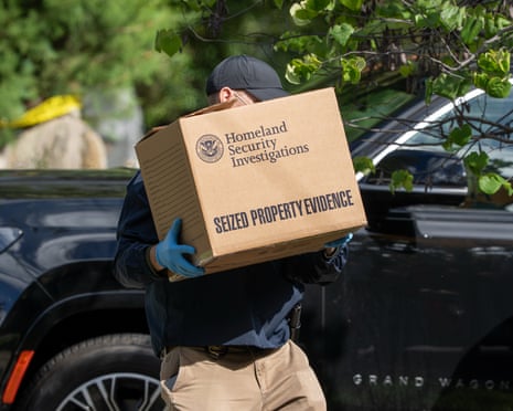 Man in baseball cap and latex gloves carries cardboard box marked 'Homeland security investigations' outside by car