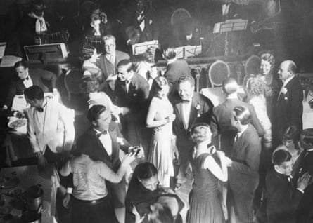 people in evening dress, the women in long gowns and men in dinner suits with bow ties, are dancing; a band, including black musicians with a banjo and saxophone, are playing