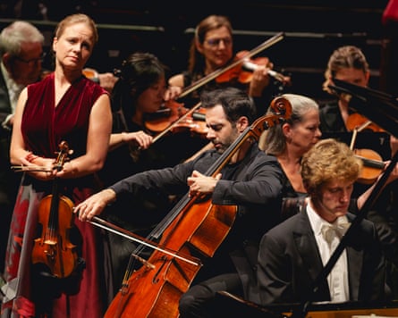 A woman in a sleeveless long dress holding a violin looks with a serious expression at a man playing a cello, with violinists seated behind them and a pianist on the cellist's right