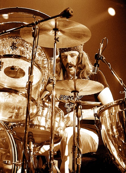 Old photo of John Bonham of Led Zeppelin, with a moustache and long hair, as seen through a drum kit