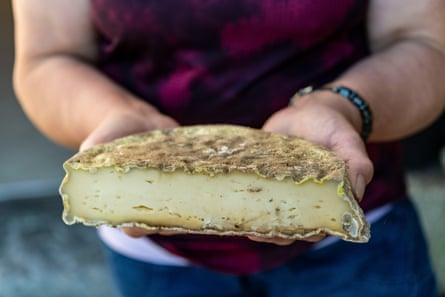A woman holds out a half-moon of artisan cheese with a thick, slightly mouldy rind and soft, creamy-looking pale yellow centre.