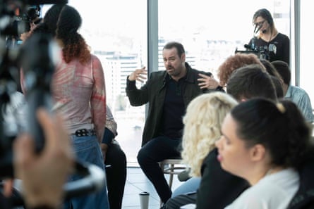 A white man with a beard and moustache sits on a chair and gesticulates
