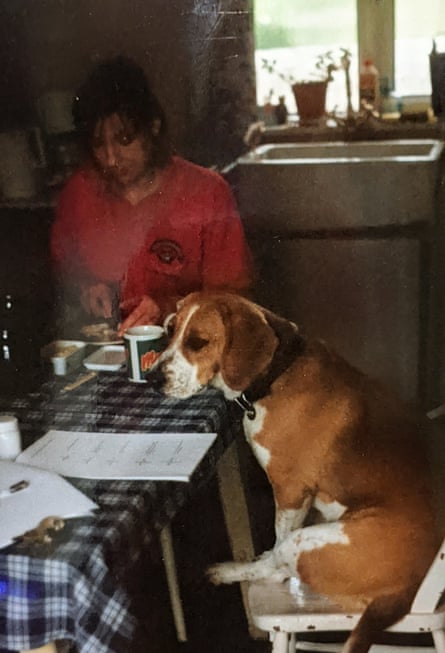 A dog sitting at a table with a checked tablecloth. A woman in a red top is at the other end of the table.
