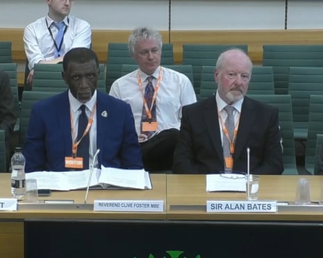 Foster and Bates seated at a desk with nameplates and microphones at a hearing, with others behind them