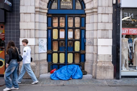 A homeless person sleeps under a sleeping bag on a doorstep as people walk by
