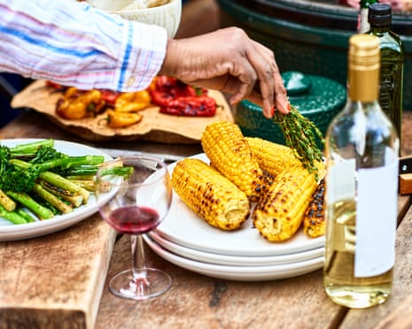 Plate of freshly barbecued corn on the cob and herbsChar grilled corn with green vegetables and wine on table, woman seasoning food with herbs, food preparation, healthy eating