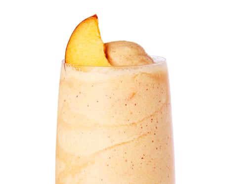 A frozen peach-colored drink in a tall stemmed glass, garnished with a peach slice