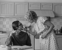 A man with slicked-back hair and glasses wearing a shirt, and a woman in a white patterned frock in an old-fashioned kitchen
