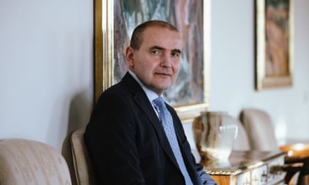 A man in a dark suit and light blue tie sitting in a room with paintings on the wall behind him
