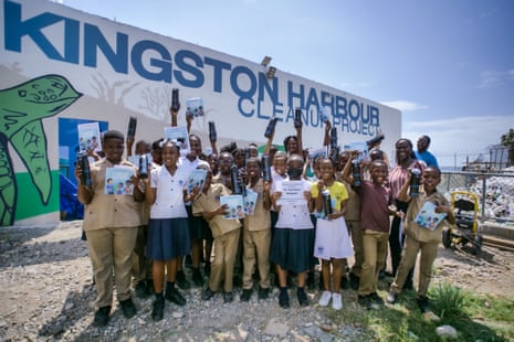 A group of about 20 Kingstonian teenage schoolchildren stand in a group holding up pictures with a huge billboard behind them referring to the Kingston Harbour Cleanup Project.