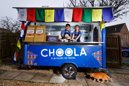 Ameer and Nicole Limbu in their blue street food kitchen-van, which houses their business called Choola. The front is hung with colourful flags and an orange cat walks in front of the van.