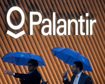 Two men holding umbrellas walk past a wall bearing the Palantir logo