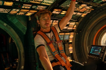 He sits in the control deck of a spaceship, wearing an orange harness